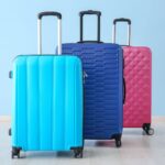Hardshell luggage in trendy teen colors and patterns against a light blue wall. Three suitcases are offset from each other, one is teal with wide vertical lines, one is navy with a horizontal line pattern and one is fuscia with a diamond pattern. All have wheels and extended handles
